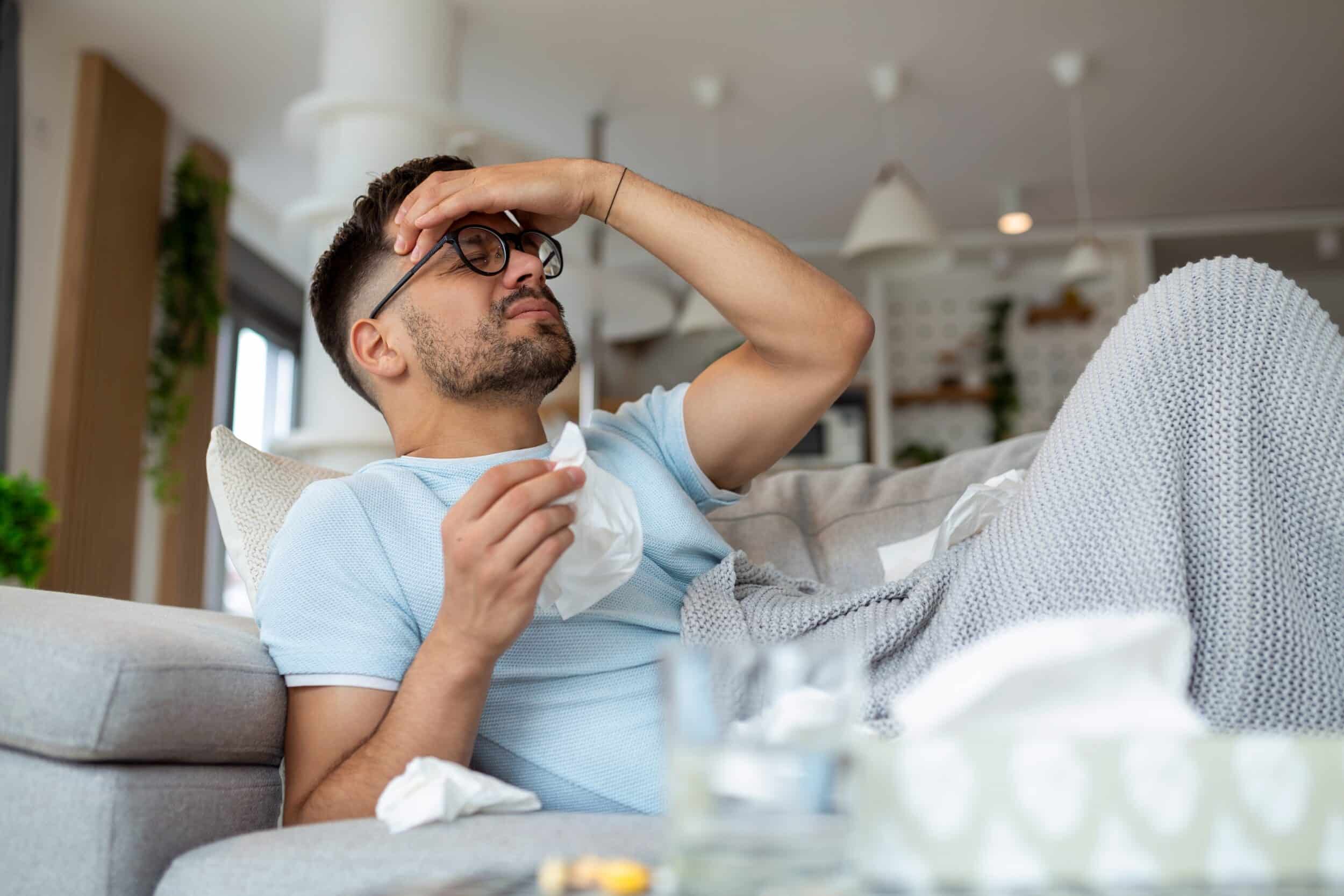 Man on sofa holding tissue and head with look of pain from sinus issues, needing Balloon Sinuplasty